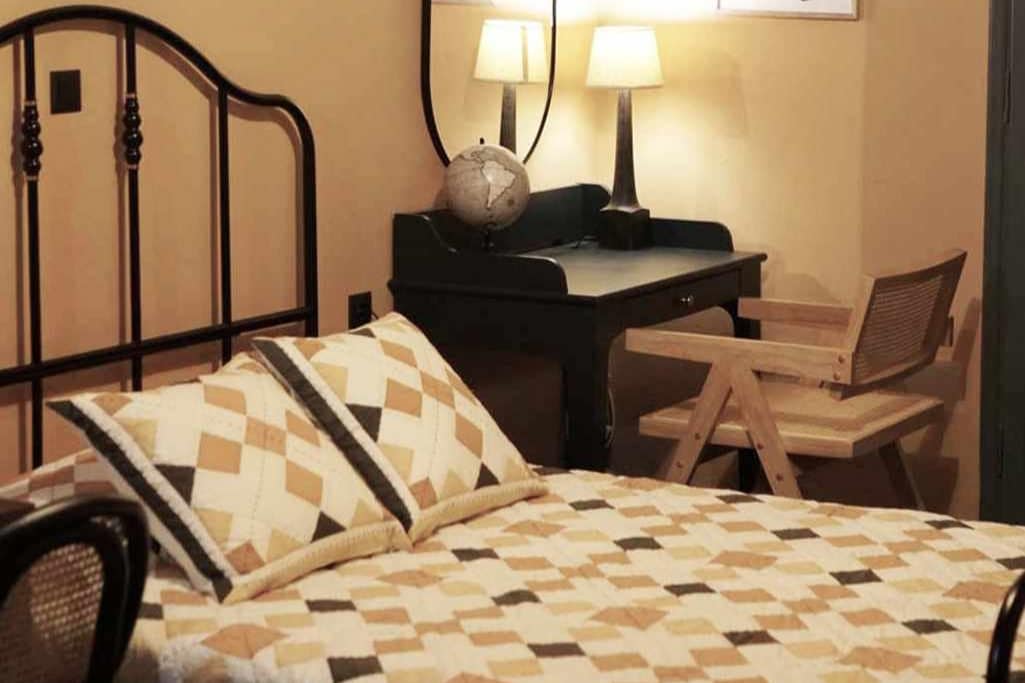 A cozy sleeping area features a quilted bedspread in warm tones. A wooden desk and a simple chair provide a functional workspace, while a bedside lamp softly illuminates the room. The globe on the desk adds a touch of curiosity to the serene ambiance.