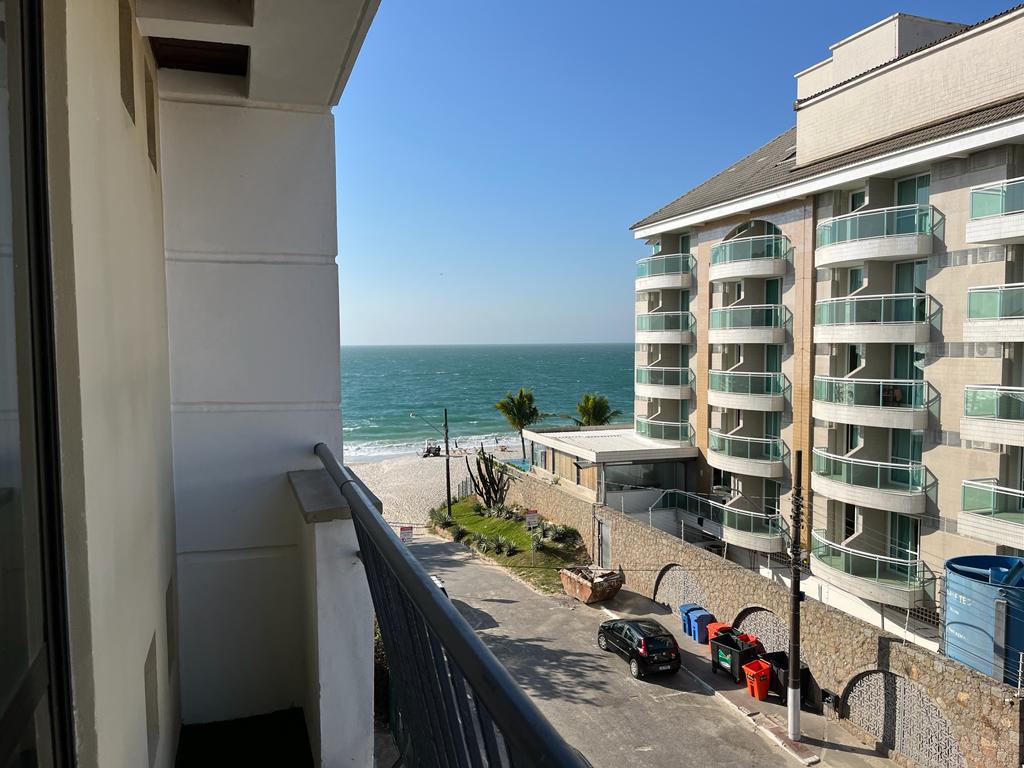 A balcony view captures the ocean's shimmering waves and sandy beach in the distance. The surrounding area includes green palm trees and a modern building structure, enhancing the coastal ambiance. Clear blue skies frame the scene, indicating a bright and inviting atmosphere.