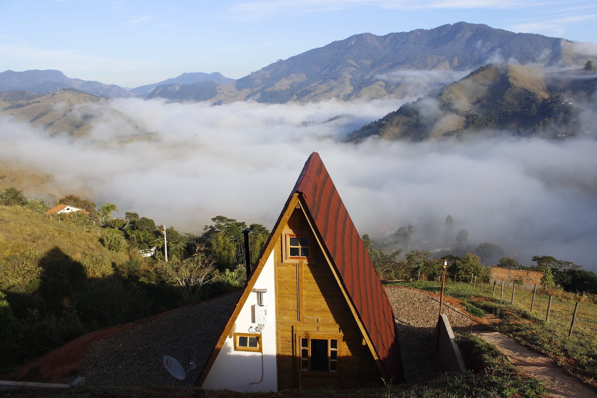 The exterior of a Swiss-style chalet is showcased against a backdrop of rolling mountains and low-lying fog. The chalet features a steeply pitched red roof and wooden walls, contrasting with the lush greenery surrounding the property.
