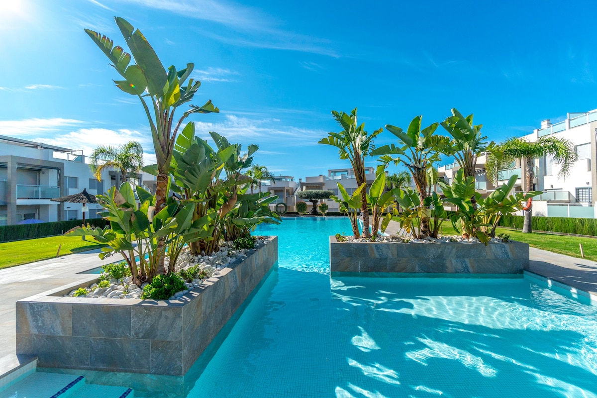 A pristine swimming pool is framed by lush, tall plants and palm trees, enhancing the sense of relaxation. Clear waters reflect the bright blue sky, while islands of greenery create inviting spaces throughout the pool area.