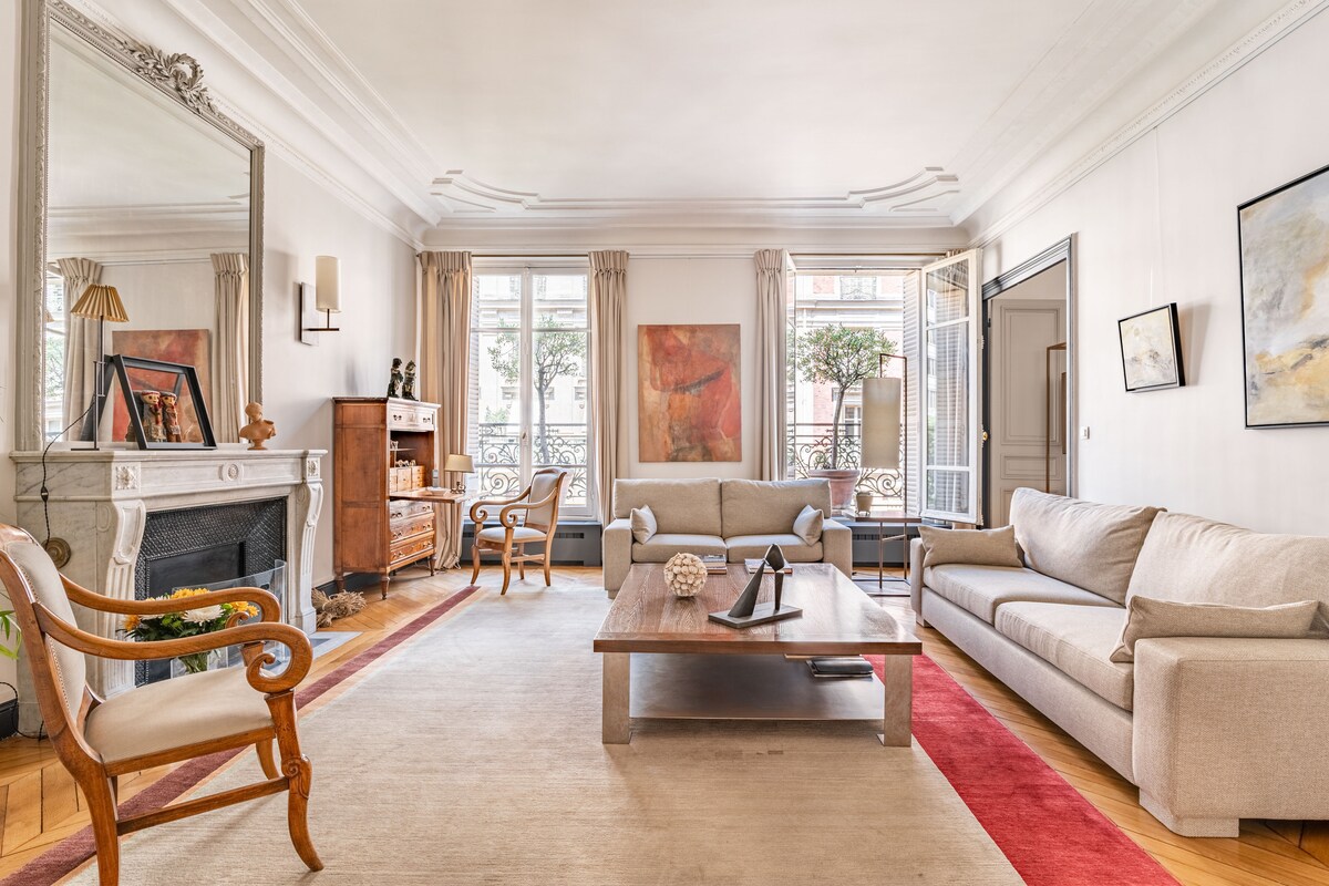 A bright living room features a mix of classic and modern design elements. Light beige sofas are arranged around a central coffee table, with a mirror reflecting the natural light from tall windows. Decorative artwork adorns the walls, and a vintage cabinet adds character.