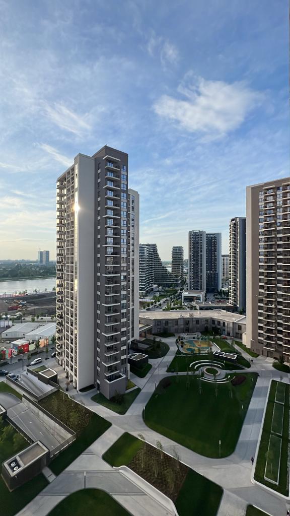 The image captures a panoramic view of a modern waterfront complex featuring multiple tall buildings. Lush green lawns and landscaped areas are visible in the foreground, while a serene river and sky occupy the background. Sunlight glints off the building surfaces, enhancing the vibrant urban atmosphere.