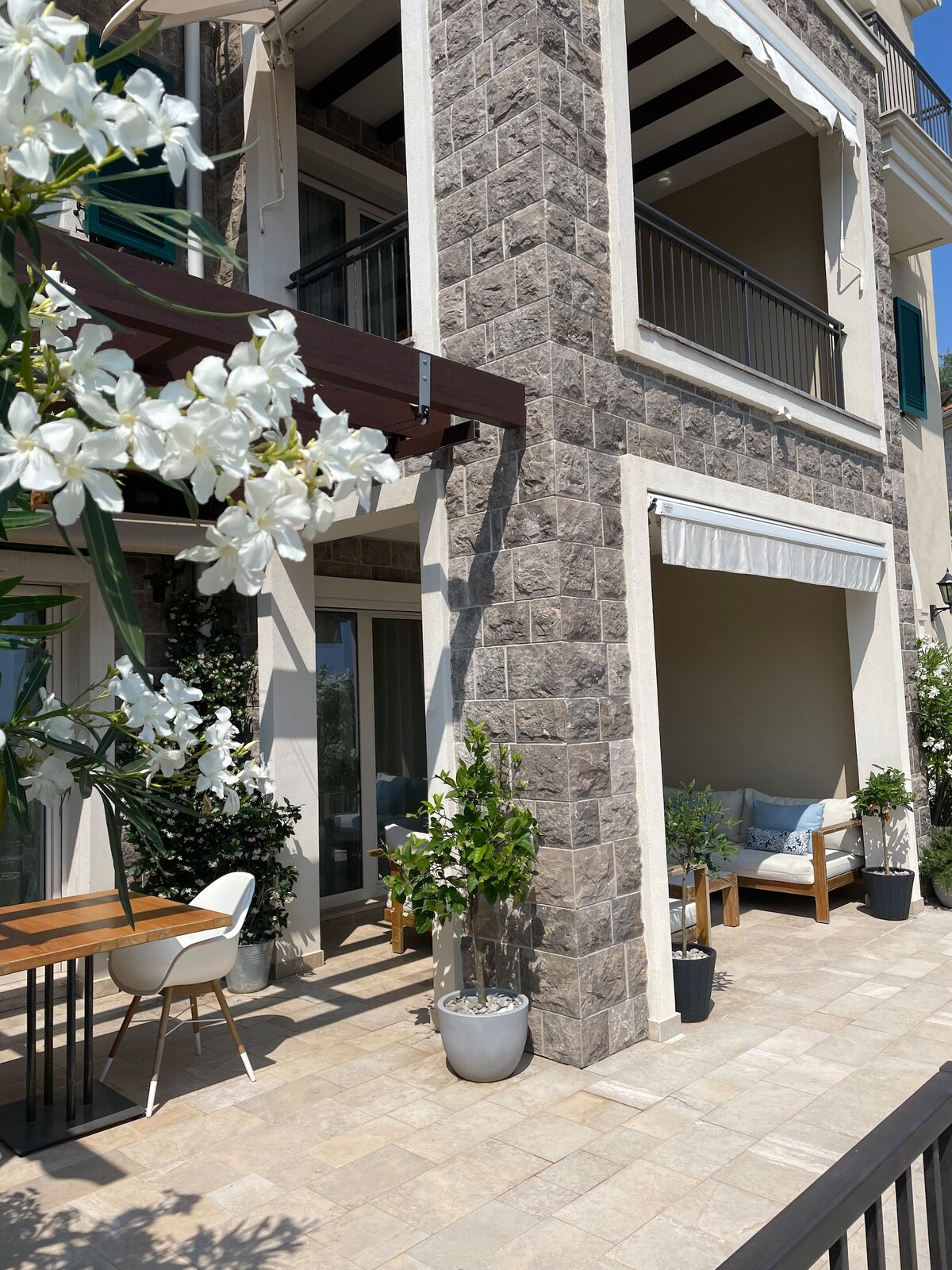 A private terrace is showcased with a light-colored stone patio, complemented by an outdoor seating area and a dining table. Potted plants and white flowers enhance the space, while large glass doors offer views into an interior living area with natural light streaming through.