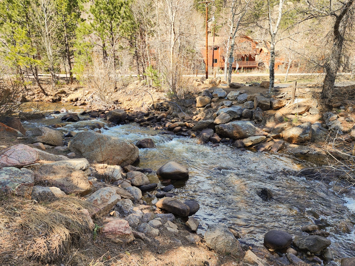 A serene stretch of the Fall River is visible, flowing over smooth stones of various shapes and sizes. Bare trees and scattered boulders frame the riverbank, while a rustic cabin is faintly seen in the distance among the trees.