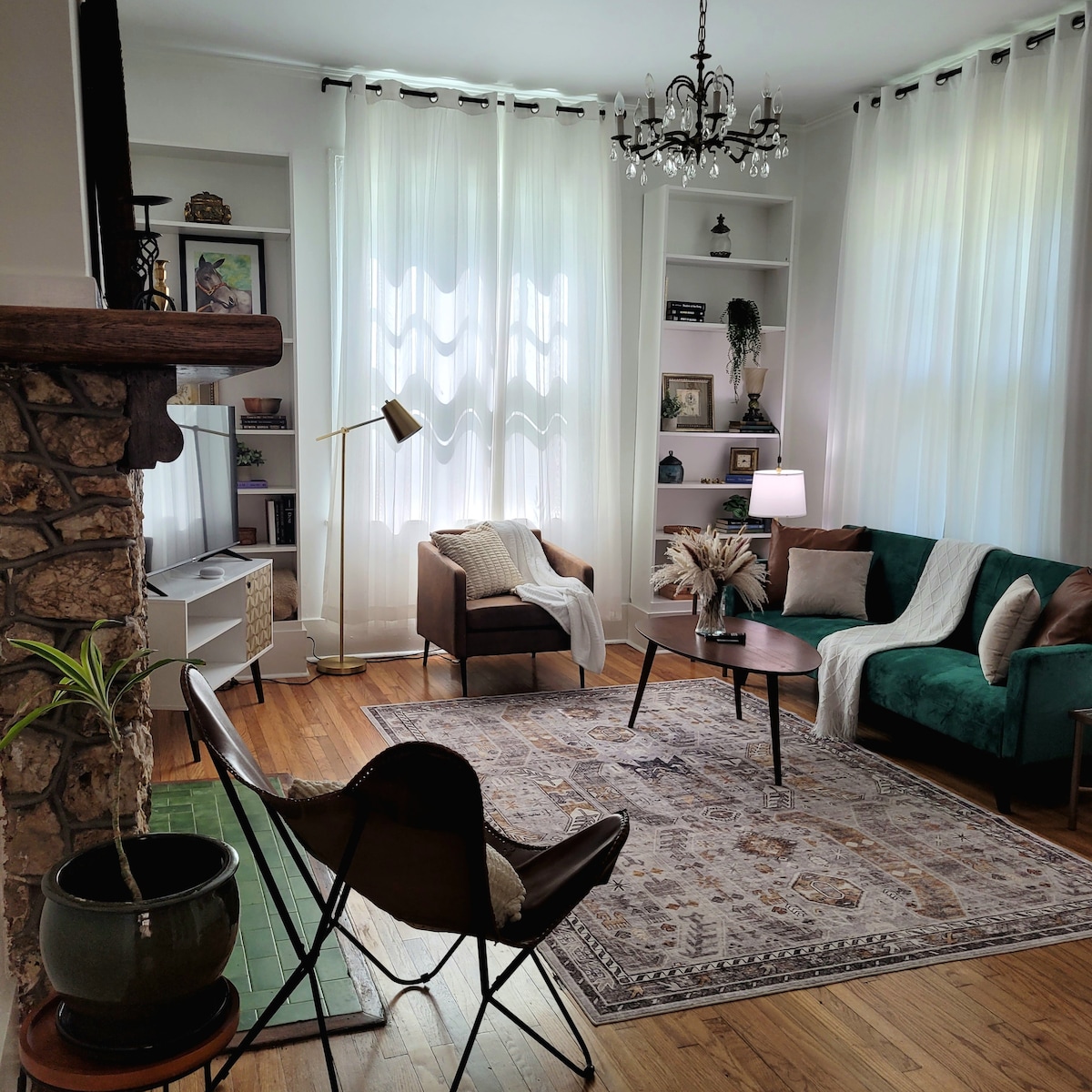 The living room features a combination of comfortable seating, including a green couch and a brown armchair. A round coffee table sits on a patterned area rug, while large windows with sheer curtains allow natural light to fill the space. A stone fireplace adds character.
