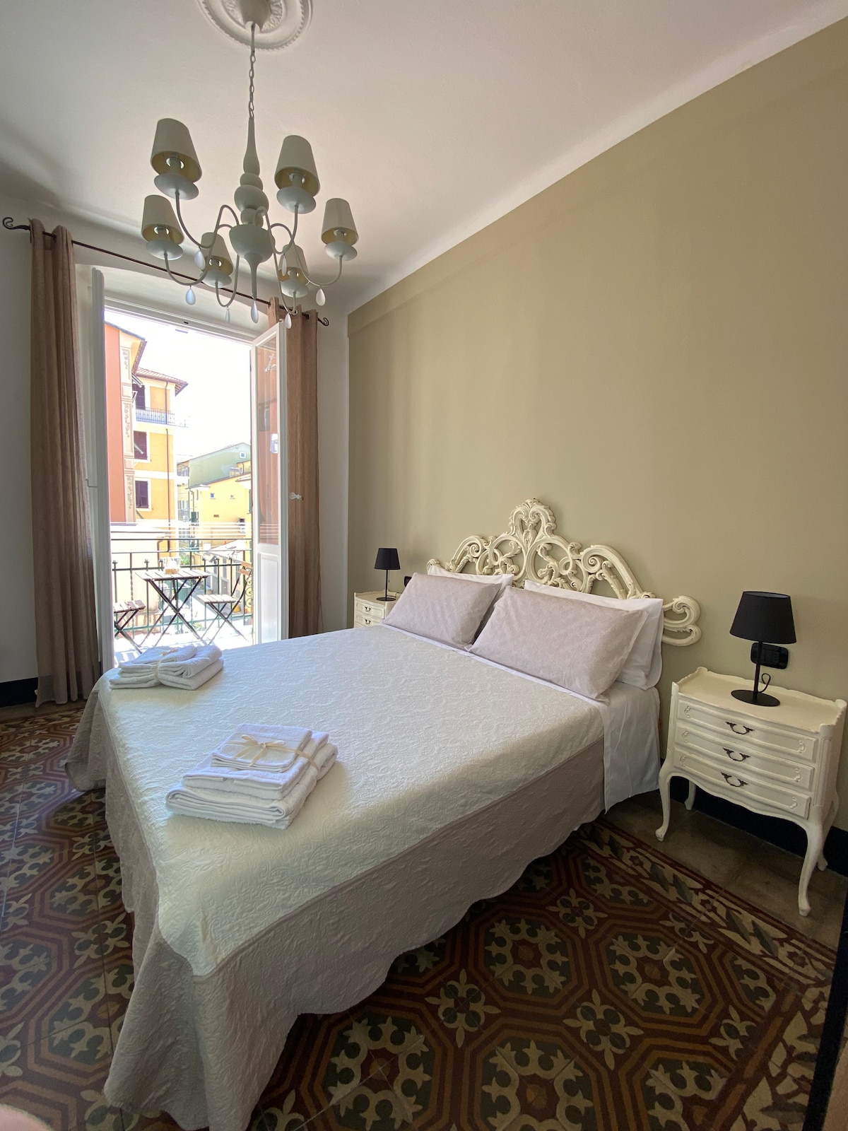A spacious bedroom includes a large bed with a light-colored quilt. Two bedside tables are positioned on either side, accompanied by black lamps. A chandelier hangs from the ceiling. Natural light enters through double doors that lead to a small balcony.