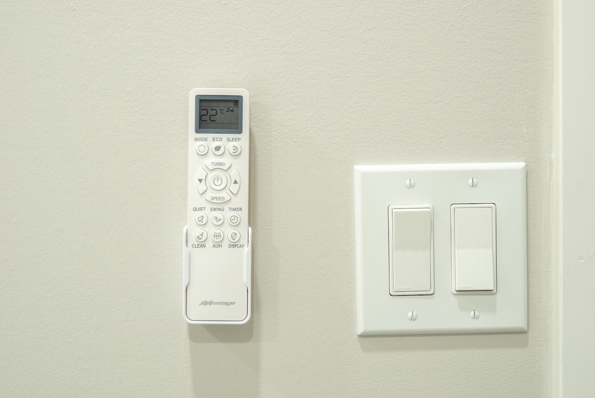 A remote control for air conditioning is mounted on a wall, displaying a temperature reading of 22 degrees Celsius. Adjacent to the remote are two light switches, positioned on a plain light-colored wall.
