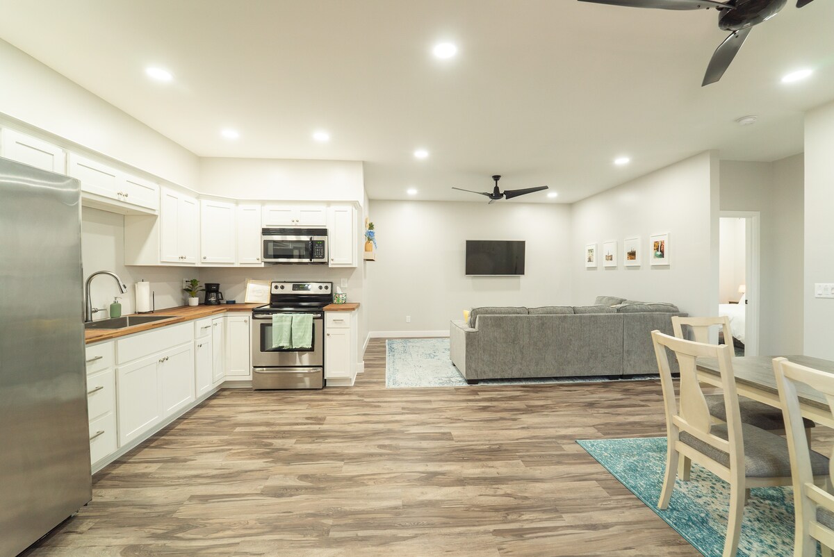 An open-concept living space includes a modern kitchen with stainless steel appliances and ample cabinetry. The dining area features a light wooden table and chairs. A spacious living area is furnished with a gray sectional sofa and a mounted television, with soft lighting enhancing the atmosphere.