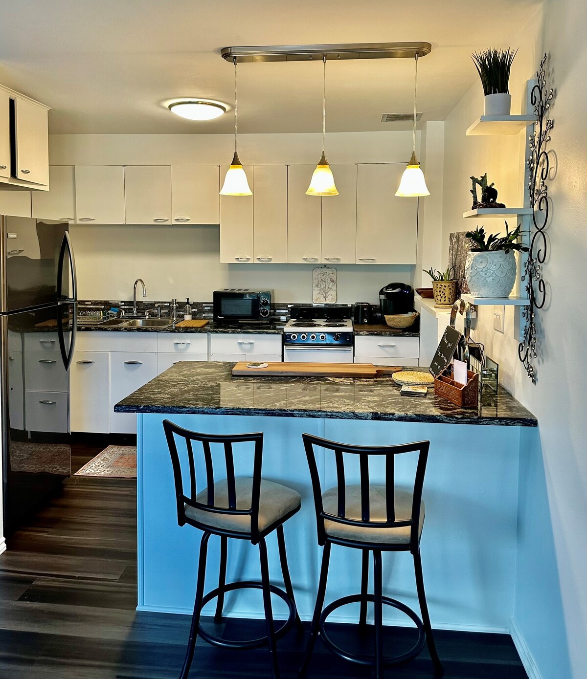 The kitchen area features modern appliances, including a refrigerator and stove. A granite countertop with two bar stools is highlighted under three soft pendant lights. White cabinetry provides ample storage, while decorative accents and cookware enhance the functionality of the space.
