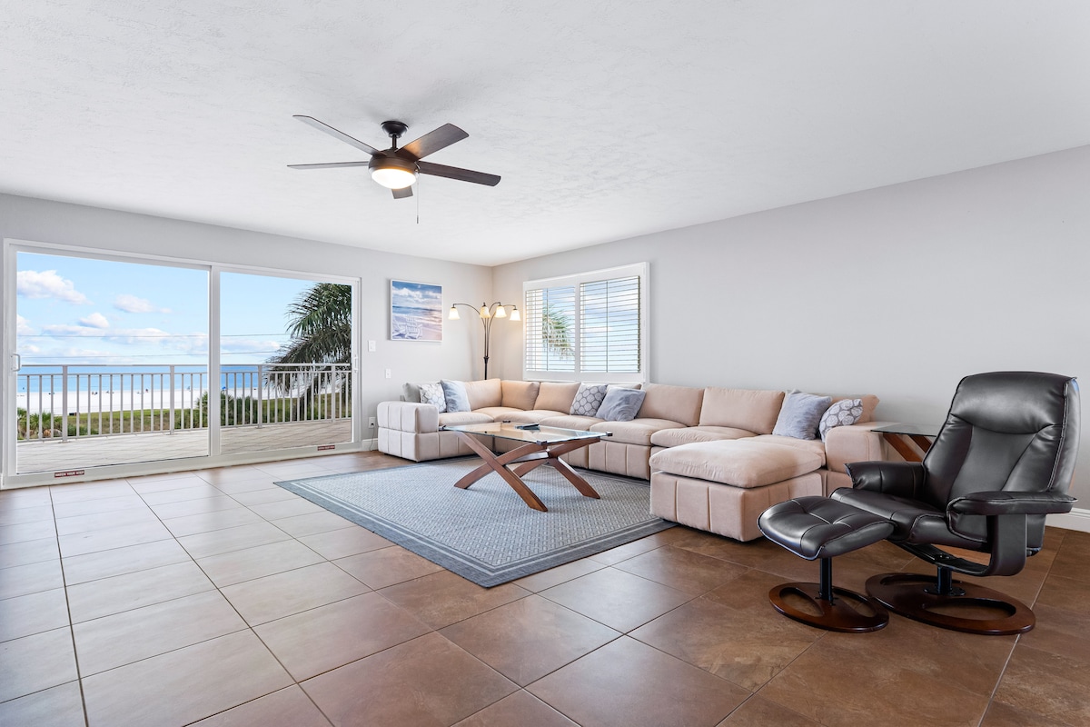 A spacious living area features a soft sectional sofa, complemented by a modern coffee table and a comfortable recliner. Large sliding glass doors provide unobstructed views of the ocean, while natural light fills the room. Neutral-toned walls enhance the sense of openness.
