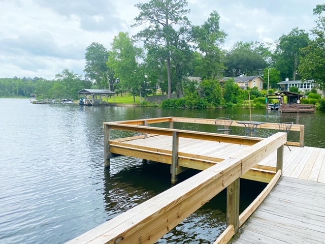 A wooden dock extends over the calm waters of Lake Talquin, surrounded by lush greenery. A pathway leads to a communal seating area with chairs, offering views of the serene lake and distant homes nestled along the shore.