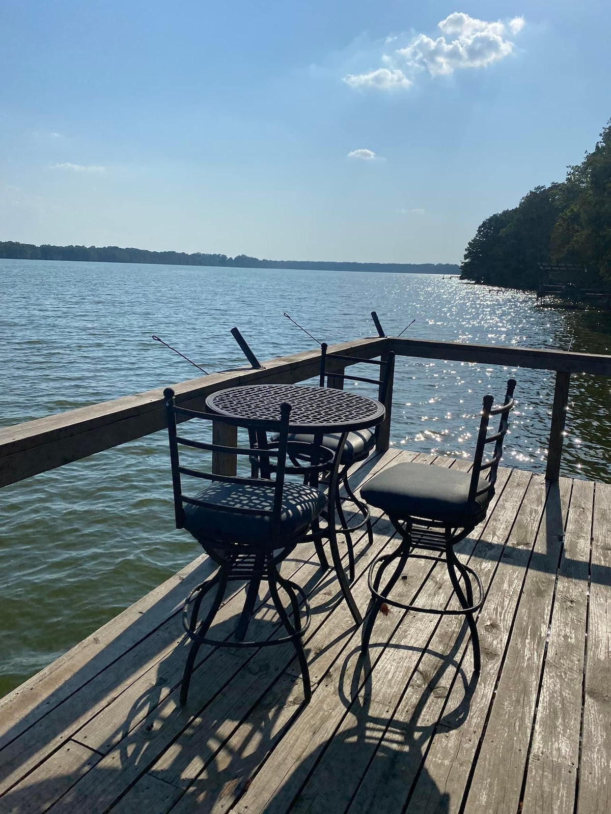 A wooden dock extends over the shimmering water, featuring a round table accompanied by two black metal chairs. Sunlight reflects off the lake, creating a serene atmosphere, with trees lining the shore in the background.