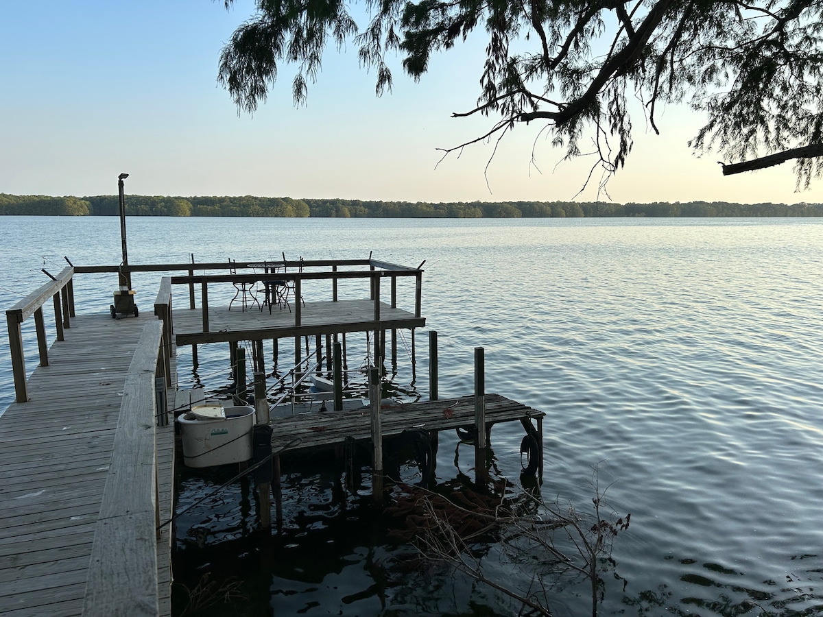 A wooden dock extends over the calm water of Lake Washington, framed by trees. A small table and chairs are located on the dock, offering a spot to relax and enjoy the serene lake views. The horizon features tree line silhouettes under a soft sky.