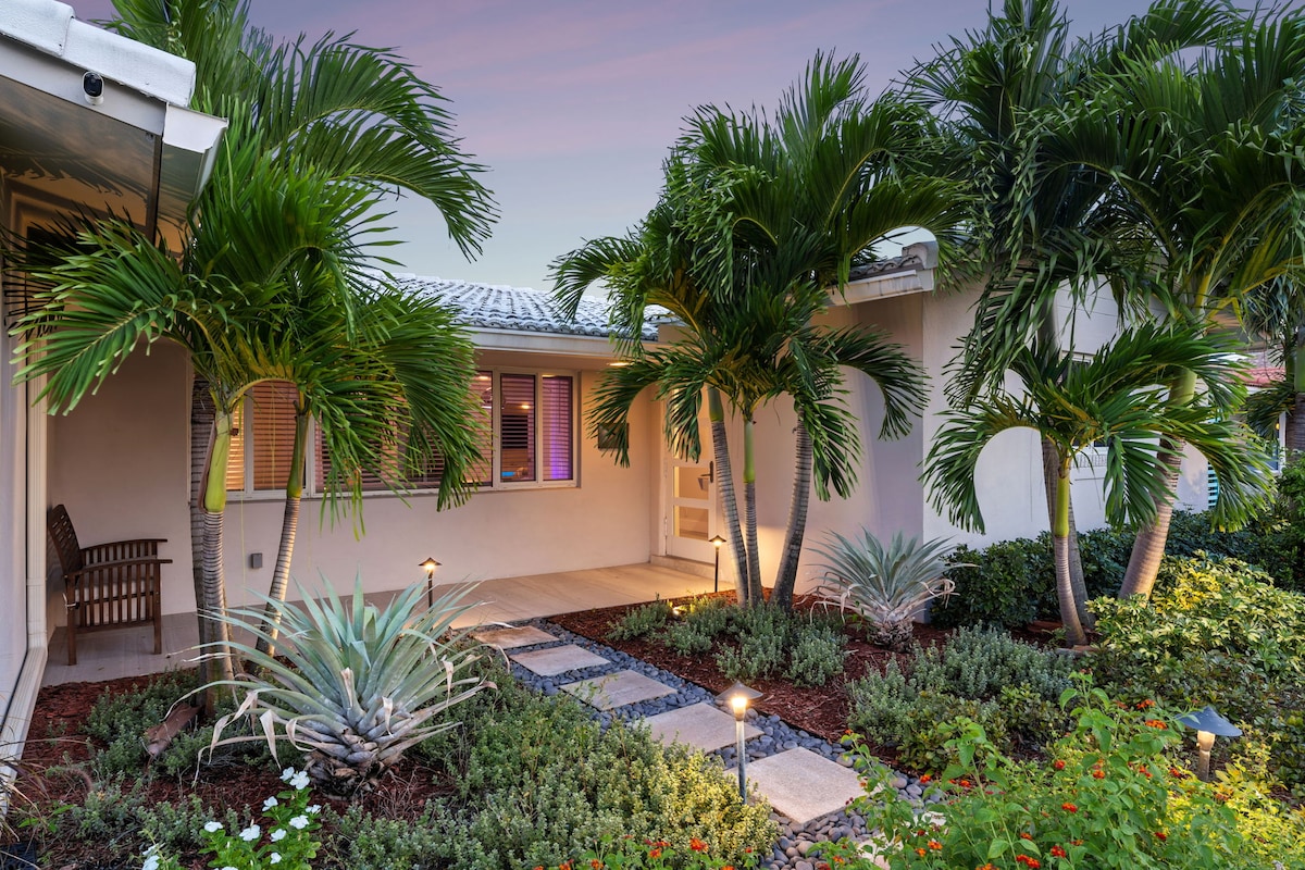 The exterior entrance showcases a landscaped pathway adorned with tropical plants and palm trees. Soft lighting highlights the stepping stones leading to the front door, while large windows invite natural light into the inviting space.