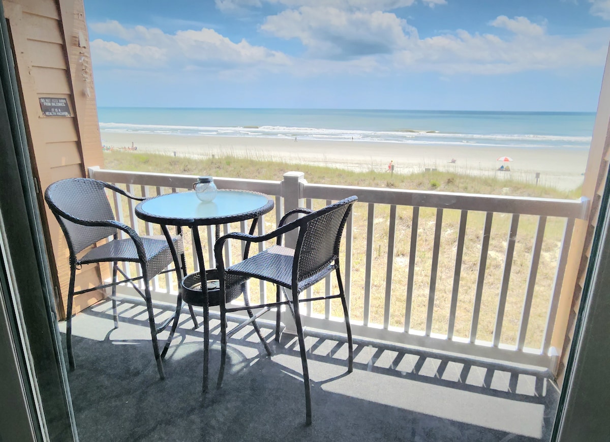 A private covered patio features a circular table surrounded by two woven chairs. An unobstructed view of the sandy beach and ocean is visible, with the shoreline stretching into the distance under a clear blue sky.