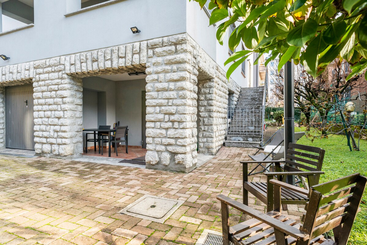 An outdoor seating area features wooden chairs arranged around a small table, set against a backdrop of stone columns supporting the building. A staircase leads to an upper level, and greenery from nearby trees adds a natural touch to the space.