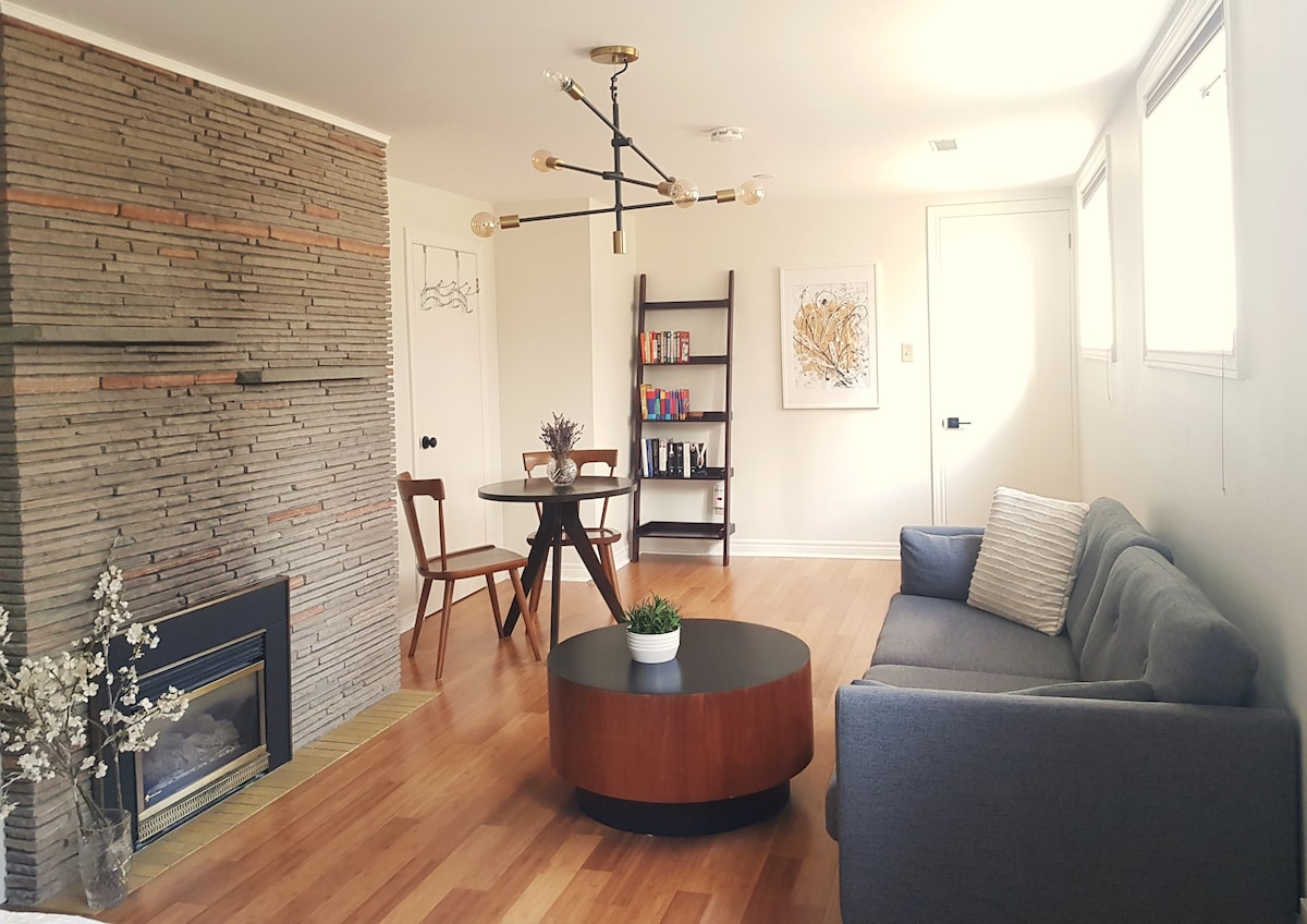 A bright living area features a large circular coffee table at its center, surrounded by a gray couch and two wooden chairs. A textured stone wall provides an interesting backdrop, and a small bookshelf holds various books. Natural light filters in through several windows.