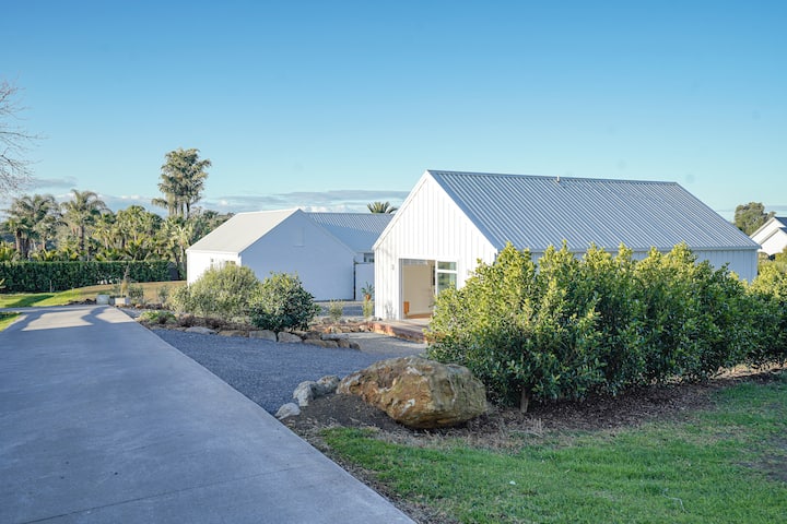 Modern white buildings with metal roofs, surrounded by lush greenery and a clear sky, with a paved driveway leading to the entrance