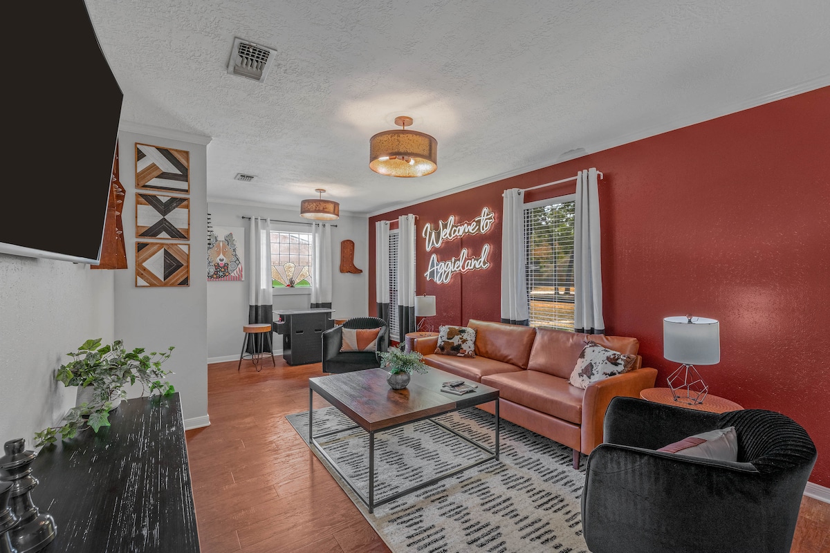 A comfortable living room showcases a combination of warm red and neutral tones. A spacious orange sofa is complemented by a patterned area rug and a stylish coffee table. Natural light enters through large windows adorned with sheer curtains, enhancing the inviting ambiance.