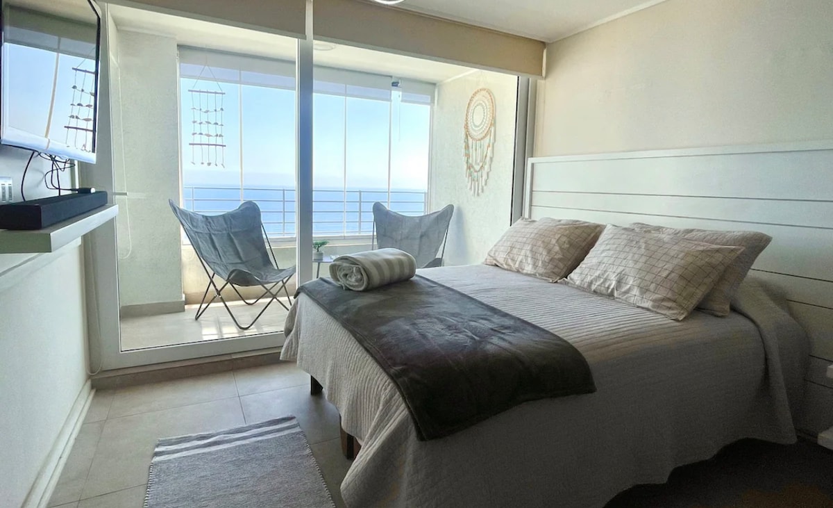 A serene bedroom is showcased, featuring a spacious bed with neutral-toned bedding. Two chairs are positioned by a large glass window, offering a clear view of the sea. Soft natural light highlights the room's calming decor and minimalist design.