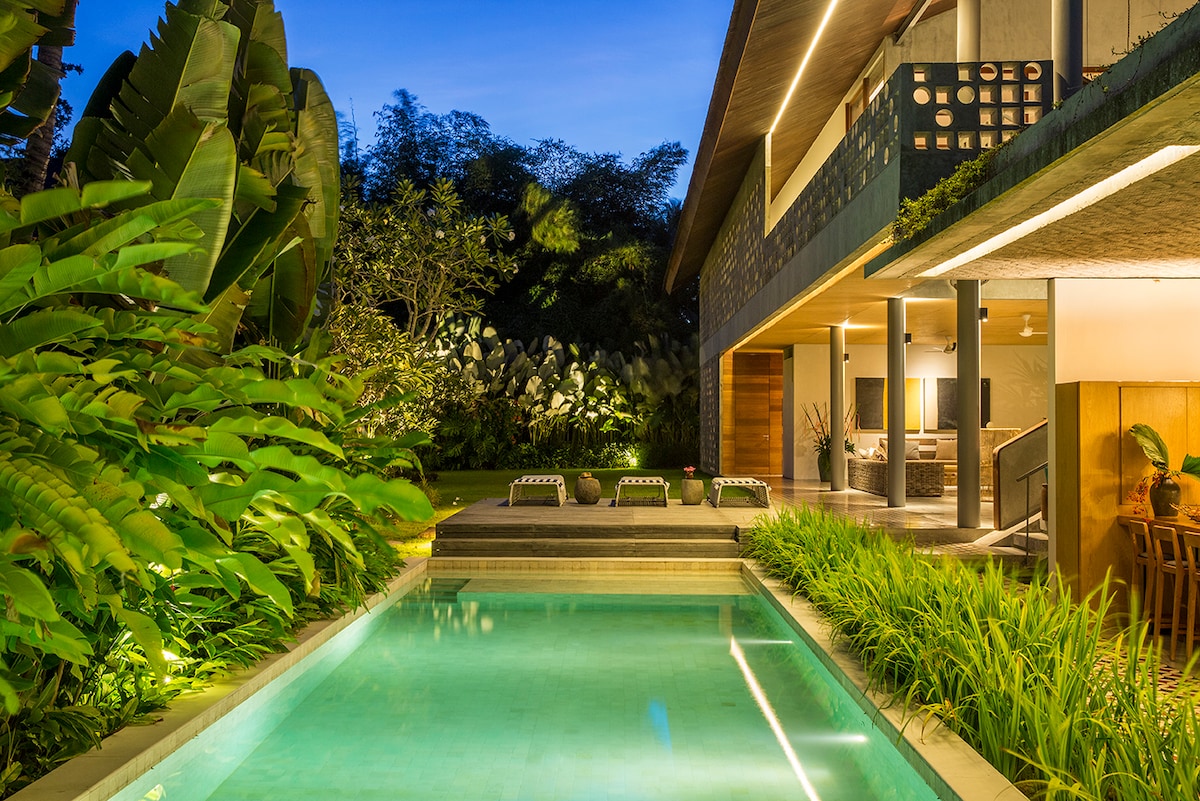 The serene swimming pool is bordered by vibrant tropical plants, creating a relaxing atmosphere. Lounge chairs are positioned near the pool, inviting moments of leisure. The villa's two-story structure is visible in the background, illuminated softly by outdoor lighting, enhancing the tranquil setting.