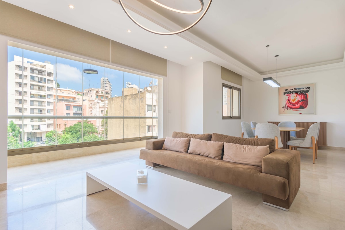 A spacious living area features large windows that offer a panoramic view of the vibrant city. A modern beige couch is paired with a minimalist white coffee table. A circular light fixture hangs from the ceiling, and a dining set is visible in the background.