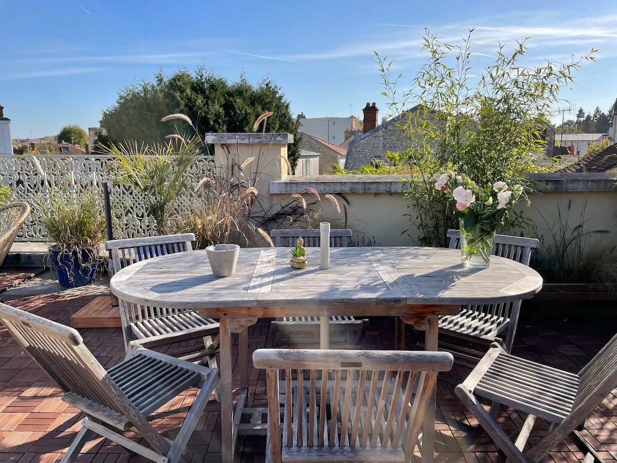 An outdoor terrace is featured, furnished with a large wooden dining table surrounded by six matching chairs. Potted plants and flowers are placed on the table, while greenery and a decorative lattice provide a serene background against a clear blue sky.