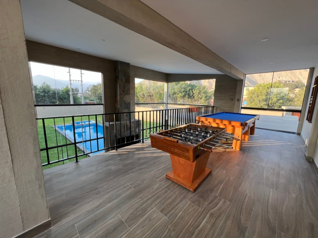 A spacious game area is shown with a table for billiards and a foosball table. Large windows provide views of the outdoor pool and green lawn, enhancing the indoor-outdoor connection. Natural light fills the room, highlighting the modern design and finishes.