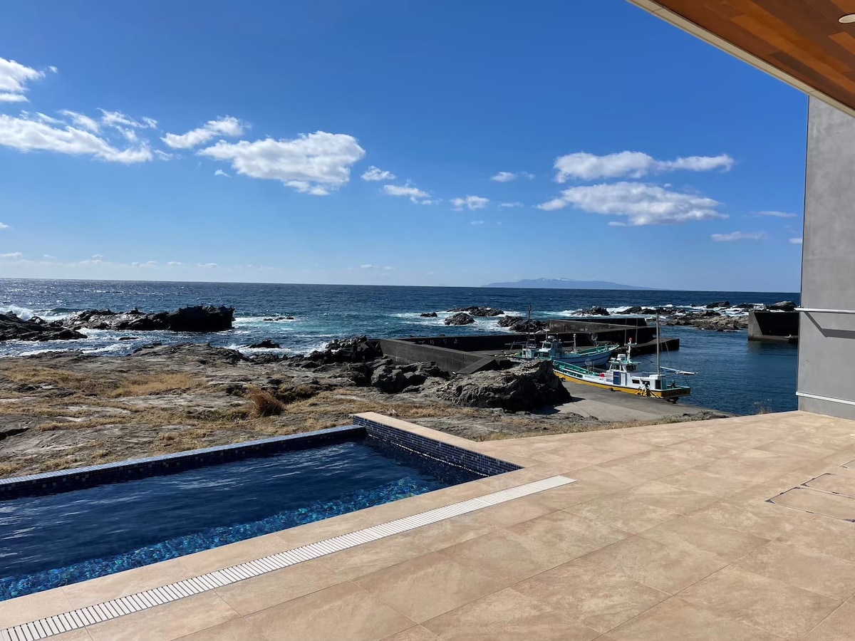 A clear view of the ocean is seen from the pool area, with rocky shorelines extending in the distance. A small boat dock is visible, adding to the coastal scenery. The pool features a sleek design, reflecting the bright blue sky.
