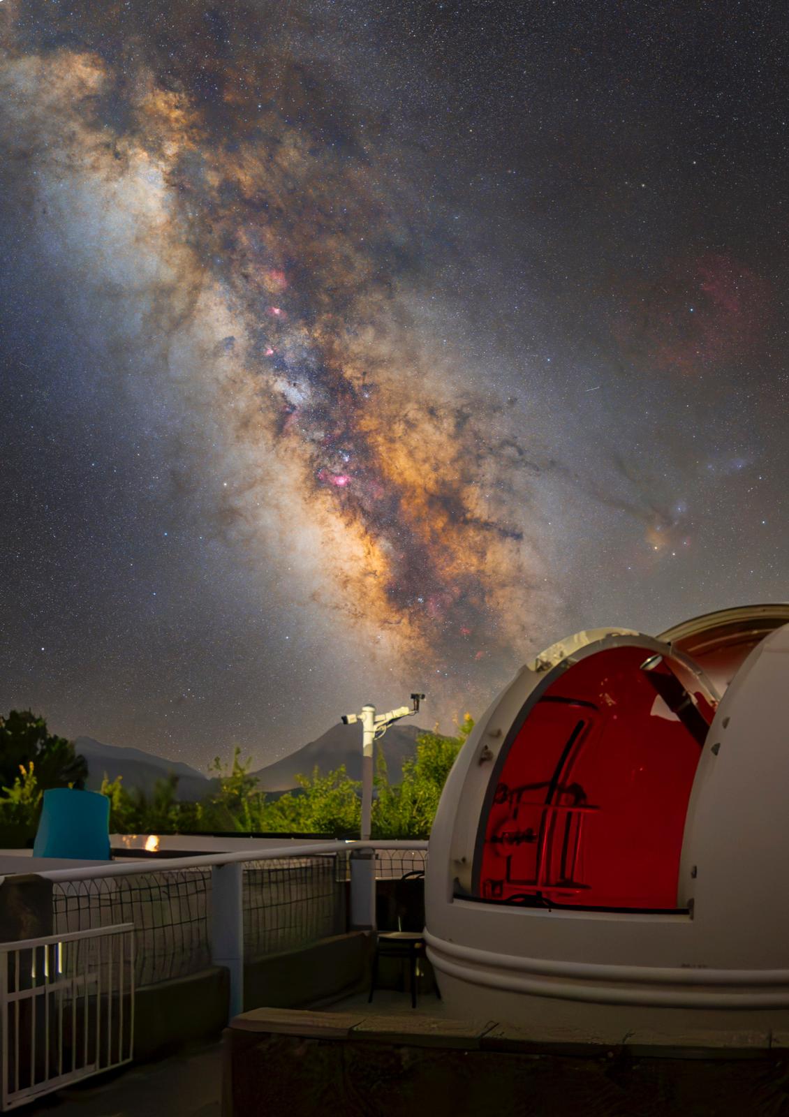 An observatory dome is illuminated against a backdrop of a star-filled sky, showcasing the Milky Way's vivid colors. The structure features a round opening, suggesting an interest in celestial observation. Trees and distant mountains are faintly visible, adding depth to the serene night landscape.