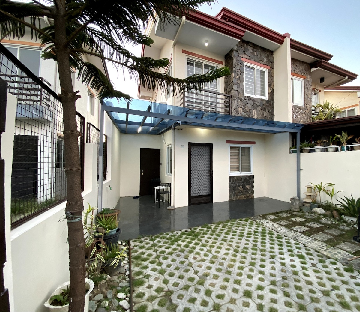 The exterior of the house features a paved patio area surrounded by plants and a decorative stone wall. A covered entrance is seen, along with a light-colored facade and window accents. The yard is neatly landscaped with geometric stone tiles and grass patches.