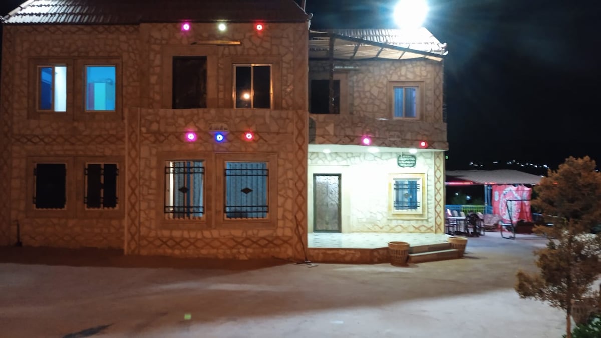 The exterior of a stone house is illuminated at night, showcasing decorative lighting around the windows and entrance. A spacious porch leads to the main door, with a gravel pathway and surrounding landscape visible. Distant gathering areas can be seen, hinting at the outdoor space.
