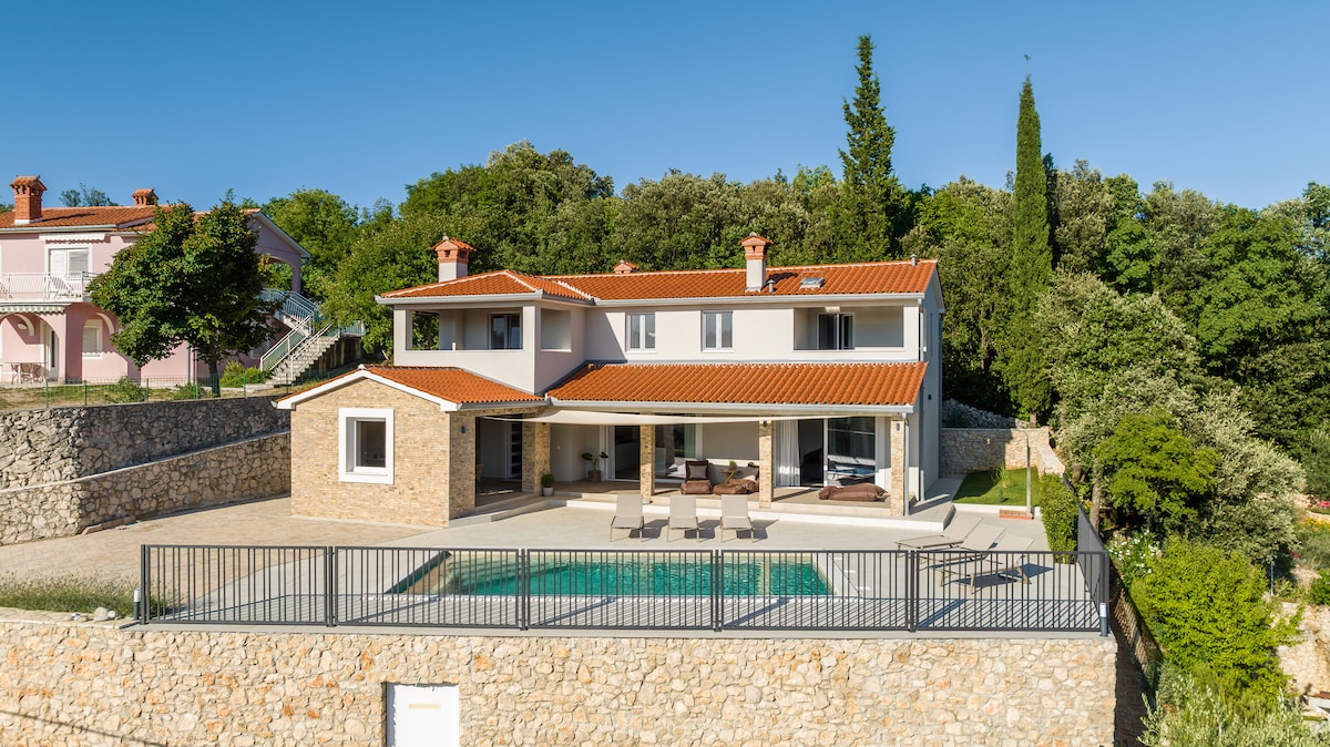 The exterior of the villa features a two-story structure with a distinct red tile roof and a combination of stone and plaster walls. A spacious terrace surrounds a private pool, accompanied by lounge chairs. Lush greenery and trees provide a natural backdrop, enhancing the serene environment.