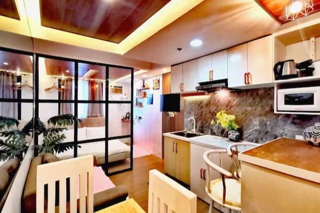 An open living space is featured with a well-equipped kitchenette, dining area, and cozy seating. A glass wall separates the sleeping area, which is adorned with greenery. Warm lighting enhances the modern design, creating a welcoming atmosphere for relaxation or dining.