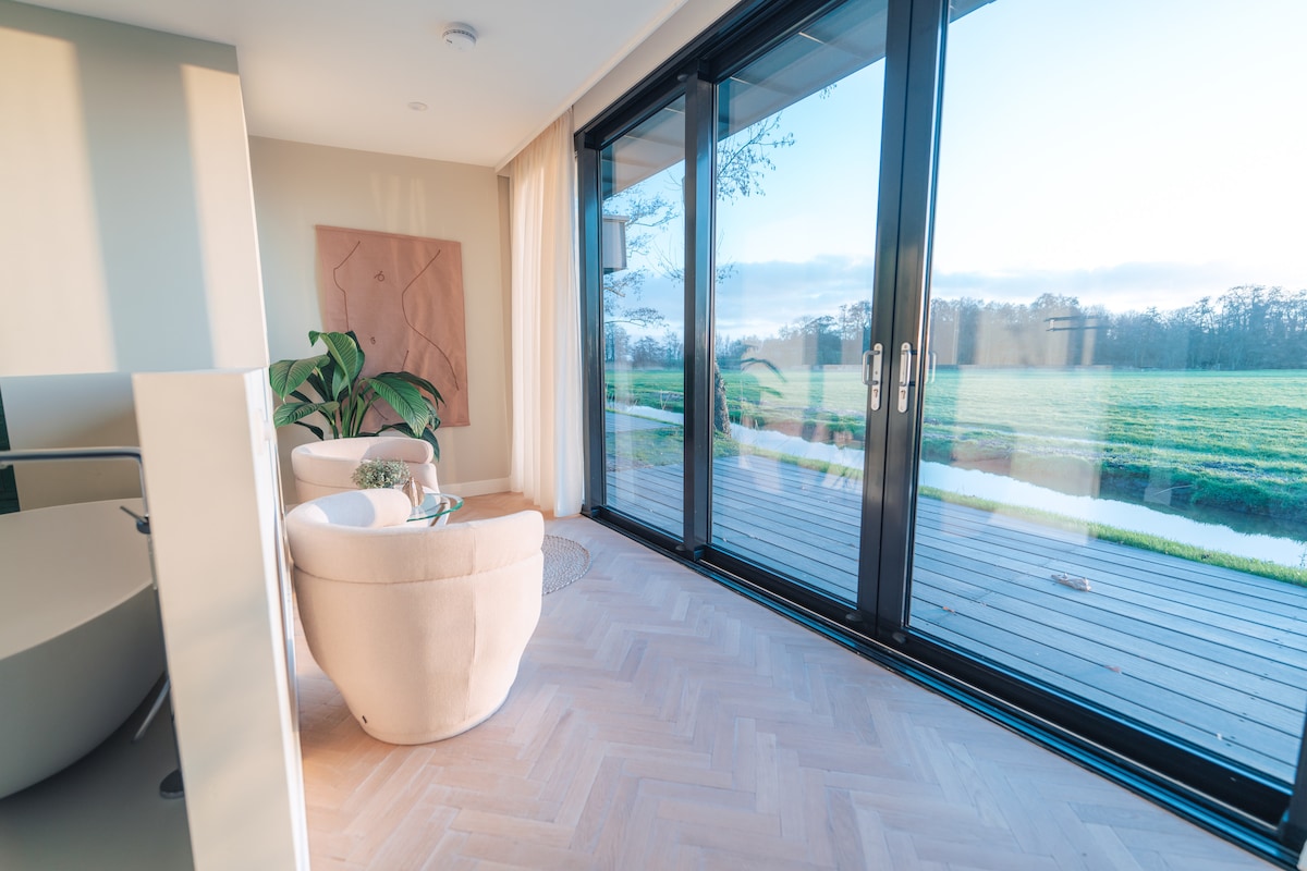 Large glass doors open to a scenic view of a lush green field. The interior features two cozy armchairs positioned beside a small table, while natural light illuminates the space through the expansive windows. A subtle plant adds a refreshing touch.