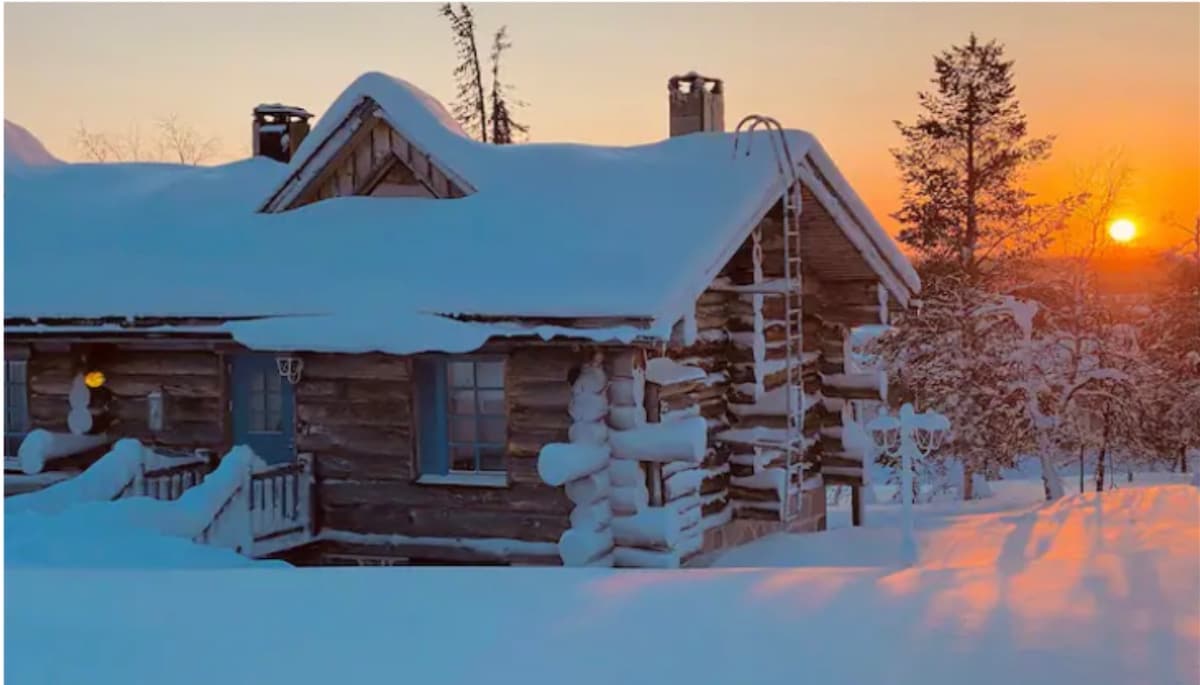 A cozy log house is surrounded by a blanket of snow, with sunlight casting warm hues across the landscape. A gentle slope leads to the front porch, while a chimney rises from the roof. Trees are visible in the background against a vibrant sunset.