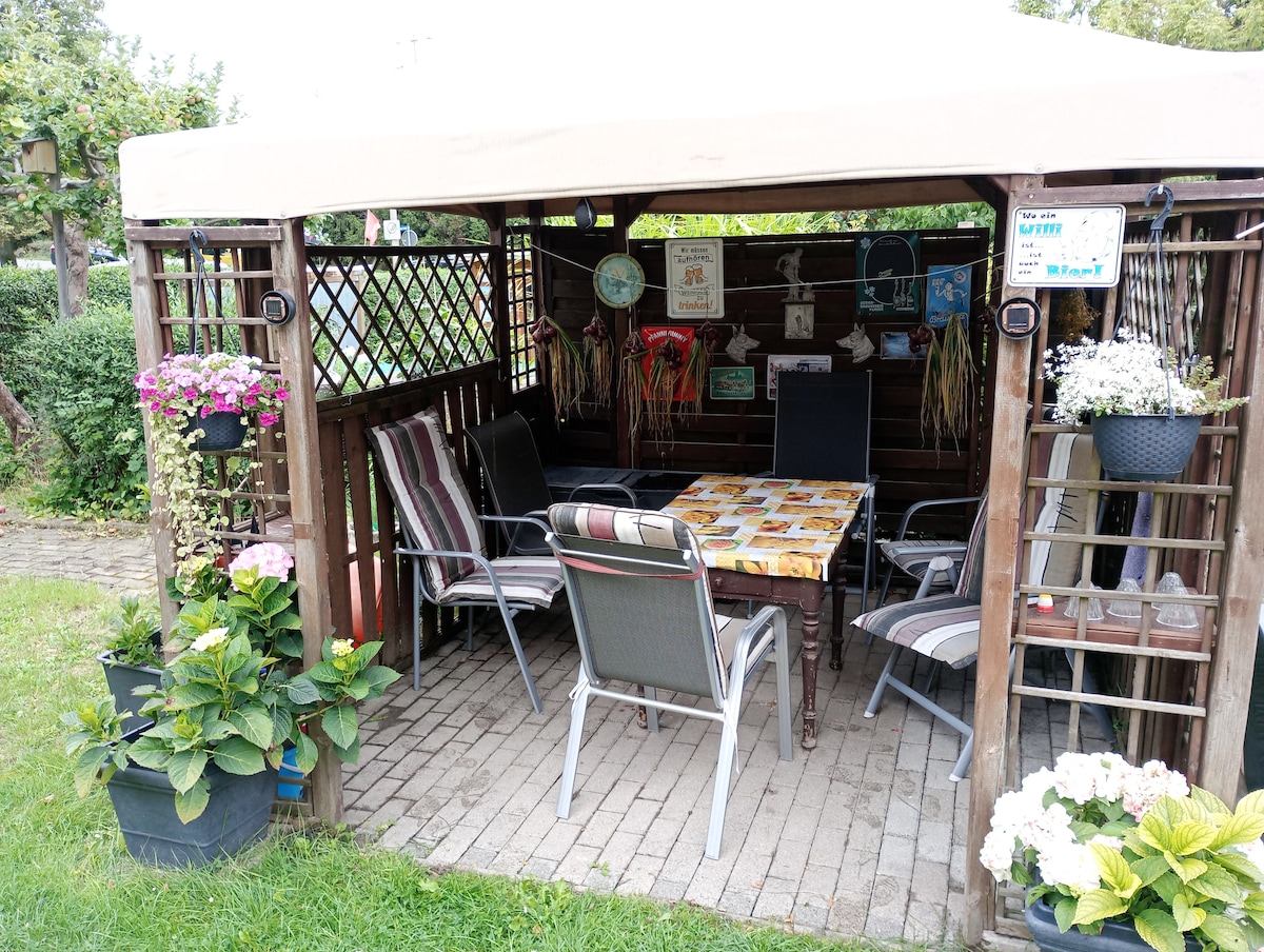 An outdoor gazebo is surrounded by greenery, featuring a table topped with a patterned cloth. Six chairs are arranged around the table, and potted flowers adorn the wooden structure. Soft lighting and decorative signs add character to the intimate seating area.