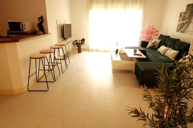 The living area is well-lit, featuring a spacious green sofa and a modern coffee table. A flat-screen TV is mounted on the wall. Four bar stools are positioned at a counter area, with decorative plants and flowers enhancing the space.