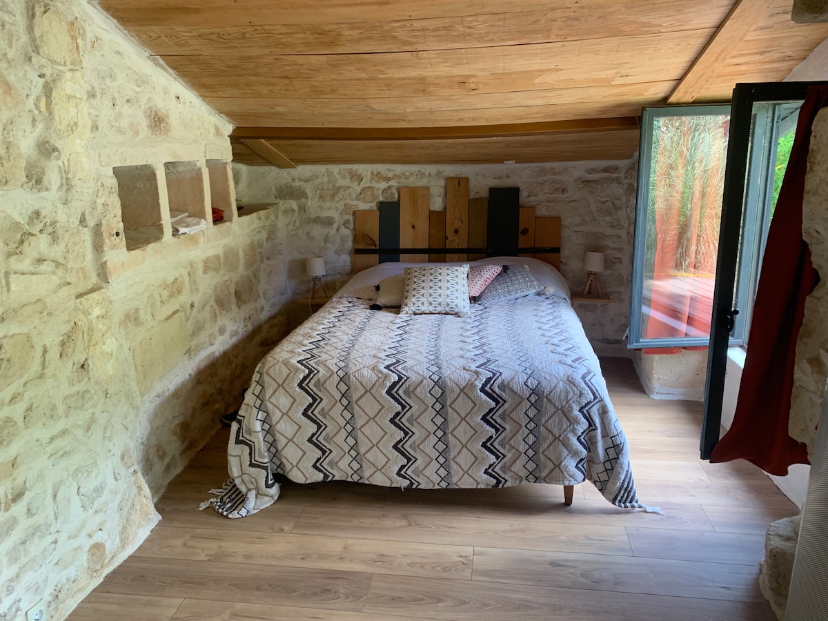 A cozy sleeping area features a spacious bed adorned with a patterned blanket. Natural wood beams and stone walls create a warm environment. A window offers a view of greenery, while illuminated bedside lamps provide soft lighting.