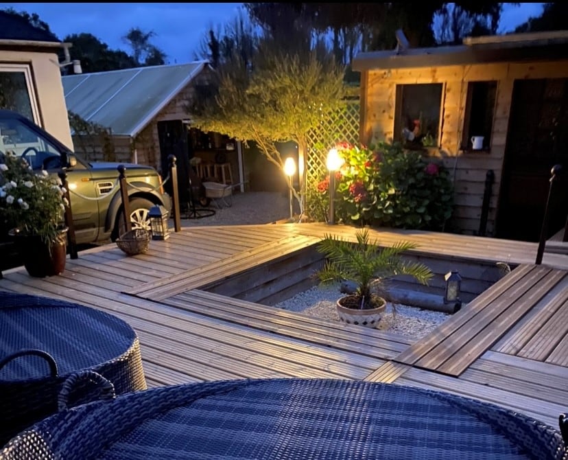 An evening view of a wooden deck area reveals two woven chairs positioned around a circular space surrounded by planked flooring. A small palm plant is placed in the center, with soft lighting illuminating the surrounding garden, enhancing the tranquil ambiance.