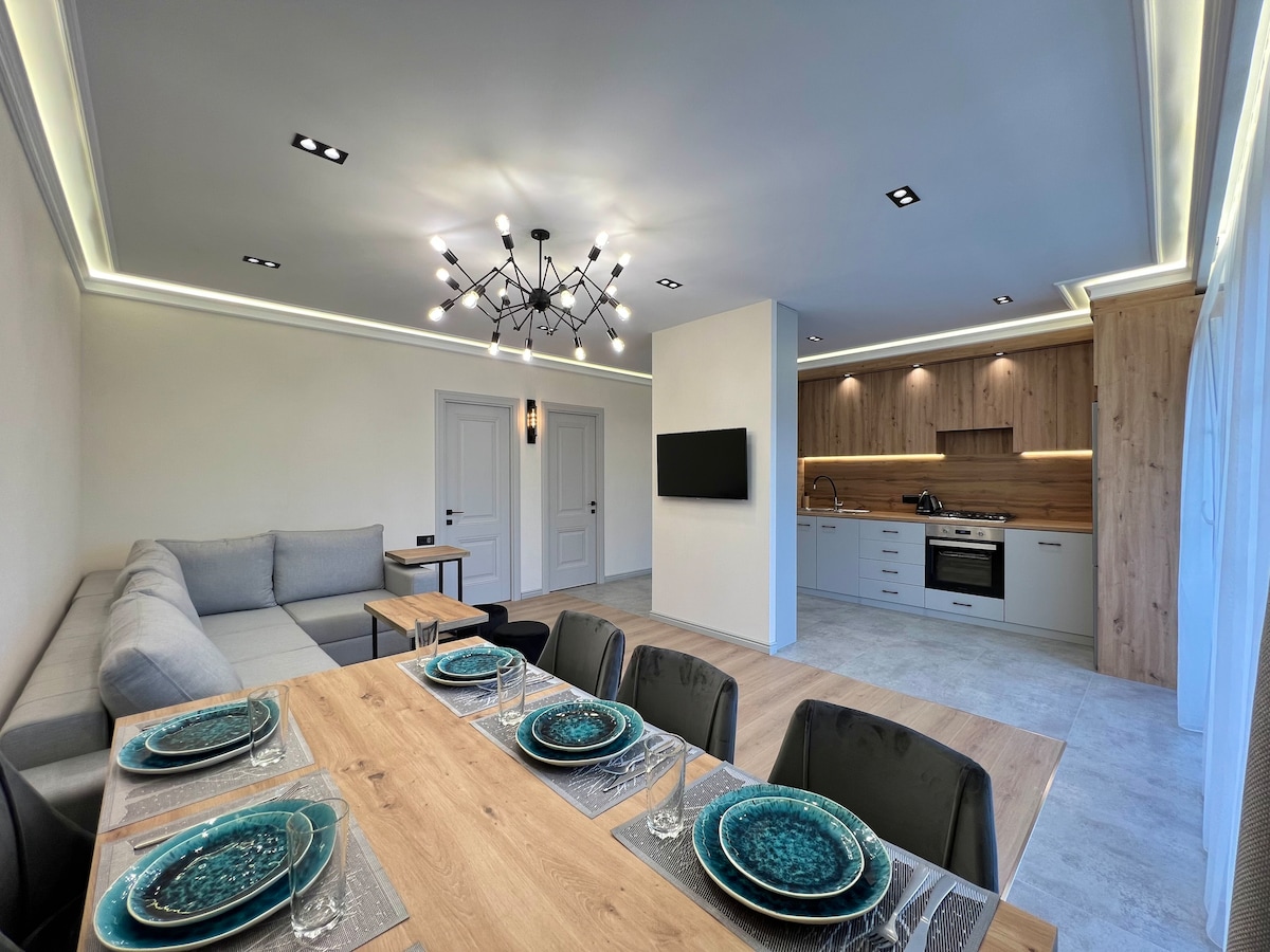 An open-plan living area features a modern kitchen with wooden cabinetry and sleek appliances. A dining table set with blue dishware is positioned beside a cozy sectional sofa. Soft lighting highlights the space, while a wall-mounted television adds functionality.