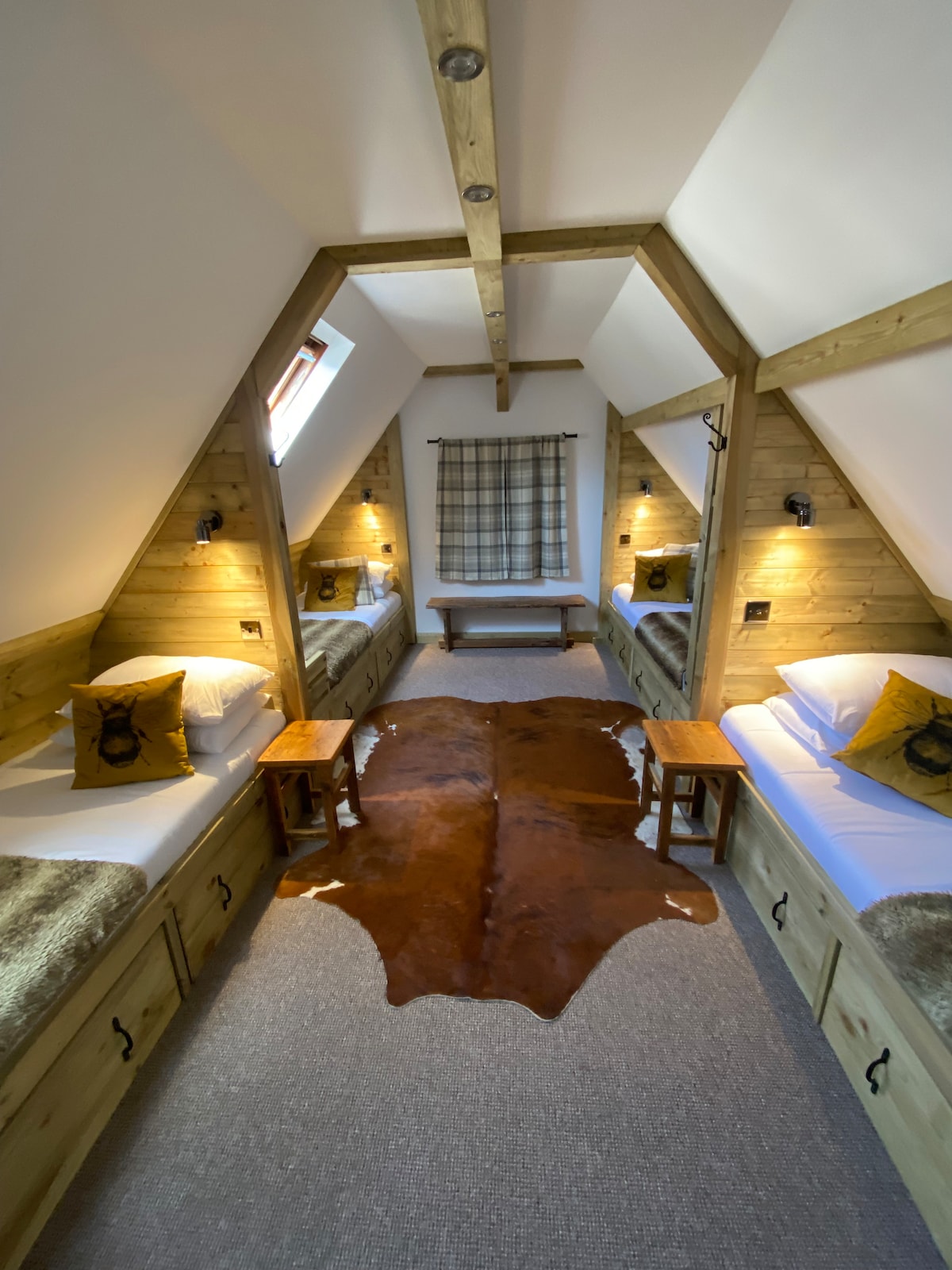 A rustic room features two single beds set against wooden paneling, with a central light brown cowhide rug on the carpeted floor. Soft, neutral accents enhance the ambiance. Two small wooden tables are placed beside each bed, and a window provides natural light filtered through a plaid curtain.