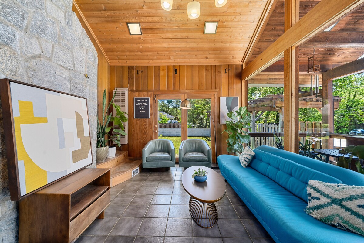 A spacious seating area features a blue sofa and two green armchairs, with a round table in the center. Large windows provide natural light and a view of the outdoors, complemented by indoor plants. A contemporary art piece is displayed on a wooden console.