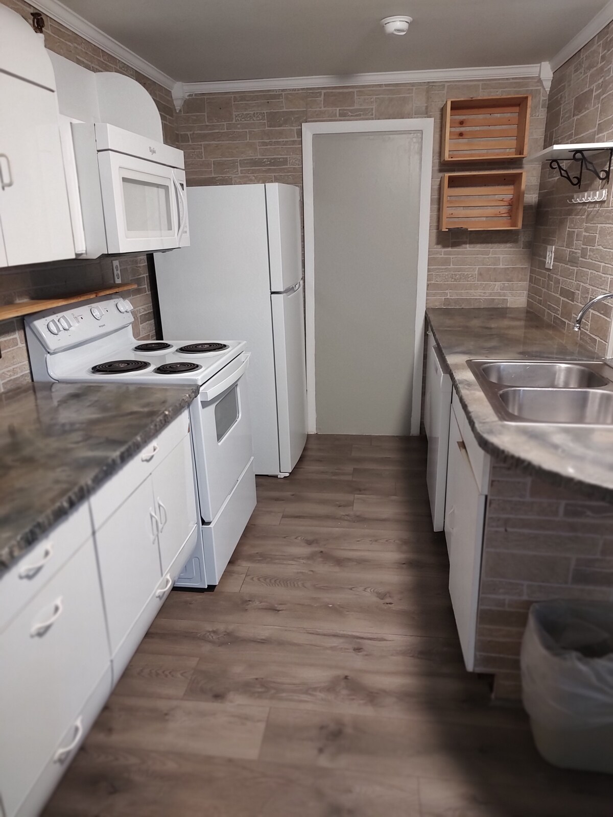 The kitchen is equipped with white cabinetry and modern appliances, including a refrigerator, stove, and microwave. A stone-patterned backsplash complements the countertops, while a double sink is positioned next to a window. Wooden shelves and a hanging rack provide additional storage options.