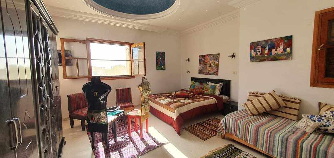 A vintage-style room features a spacious bed, a separate single bed, and a decorative mannequin. Colorful artwork adorns the white walls, and natural light enters through multiple windows. A patterned rug covers the floor, enhancing the room's unique atmosphere.