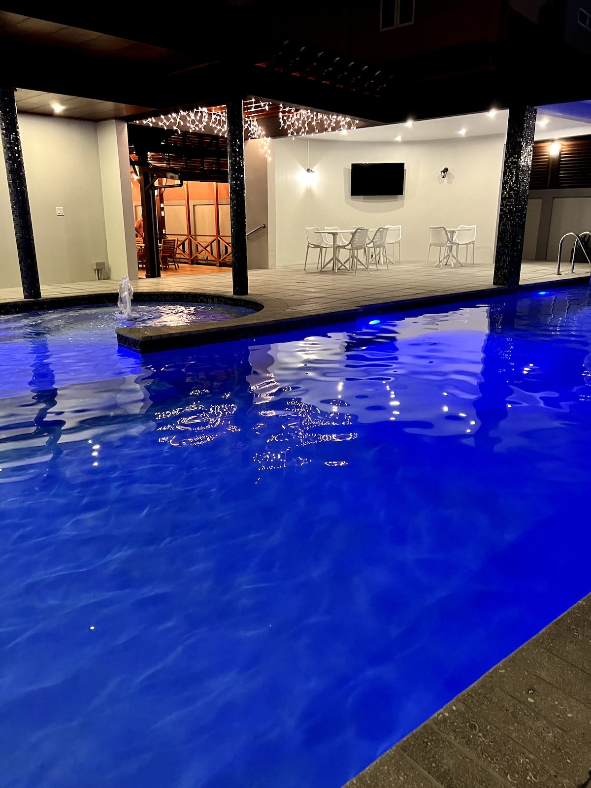 A serene pool area is presented, featuring illuminated blue waters and a gently flowing fountain. The elegant seating area under soft lighting is visible in the background, complemented by a wall-mounted television. The outdoor space offers a peaceful setting for relaxation.