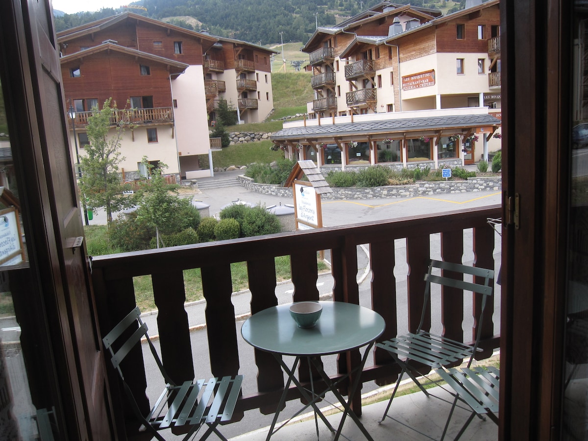 A balcony view showcases a small round table with a bowl resting on it, accompanied by two folding chairs. The scene includes neighboring wooden buildings and greenery, set against a backdrop of mountain slopes. The entrance to a nearby establishment is also visible.