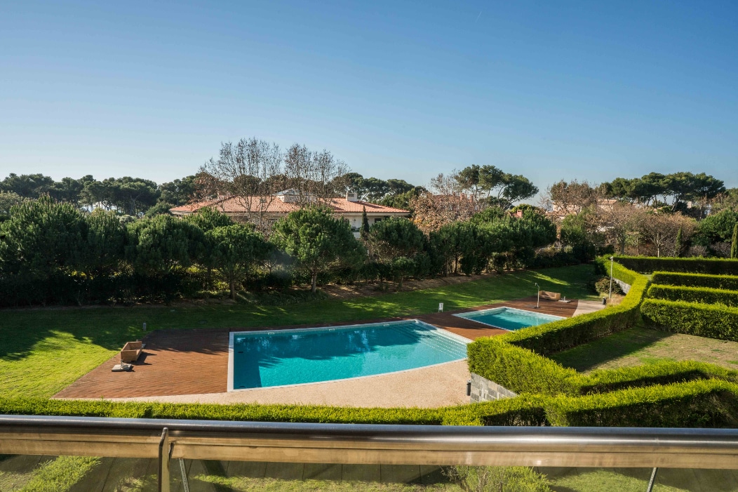 A serene outdoor area is showcased, featuring a well-maintained swimming pool surrounded by neatly trimmed hedges and green grass. Sun loungers are visible on the deck, providing a tranquil space for relaxation. The landscape is framed by tall trees in the background.