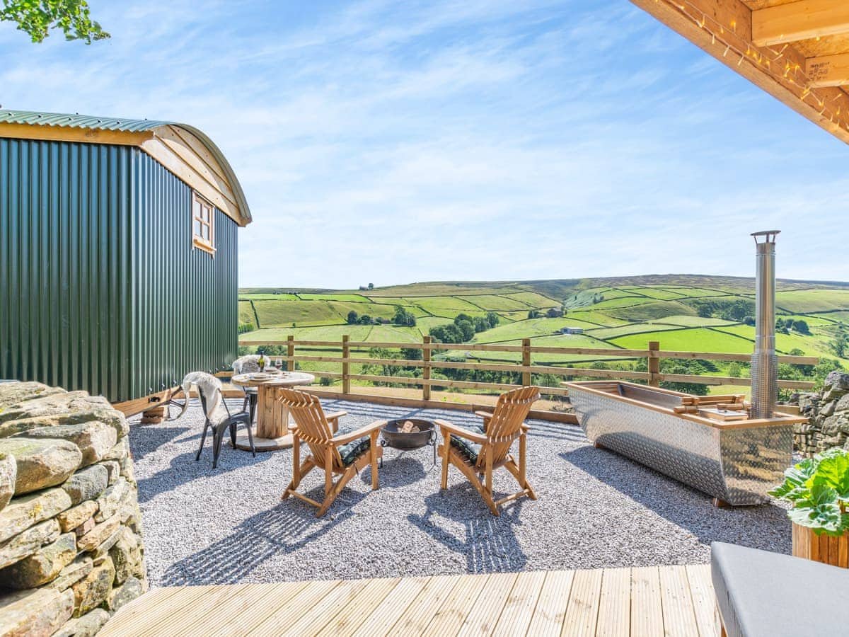 An outdoor seating area features two wooden chairs and a bench surrounding a central fire pit. The space is set on gravel, offering expansive views of the surrounding green hills and valleys. A private hot tub is positioned nearby, adding to the tranquil ambiance.