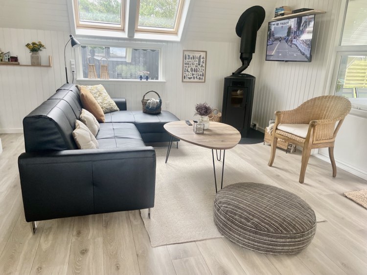 A bright living area features a modern black sectional sofa paired with a round coffee table. A cozy armchair and a soft floor cushion enhance the comfort. Natural light enters through skylights, while a flat-screen TV is mounted on the wall beside a wood-burning stove.